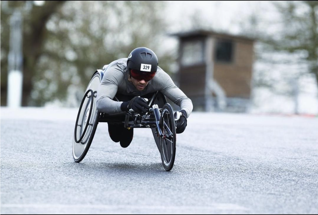 An athlete wearing a helmet races in a three-wheeled racing wheelchair on a road