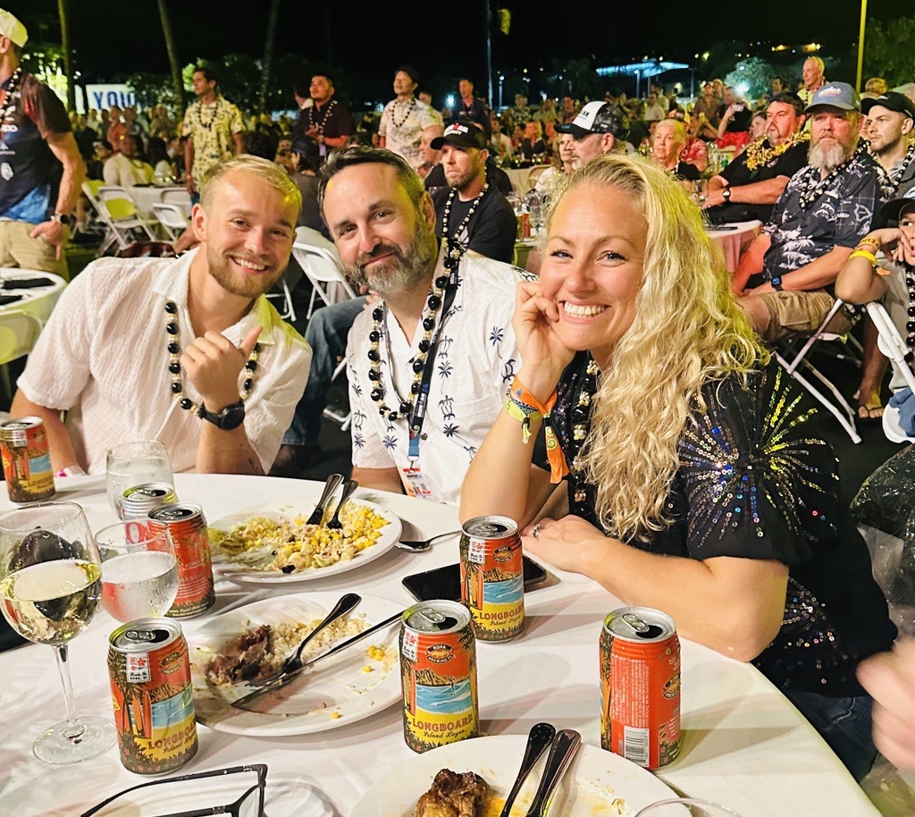 Three people smile together at a table during an outdoor evening celebration